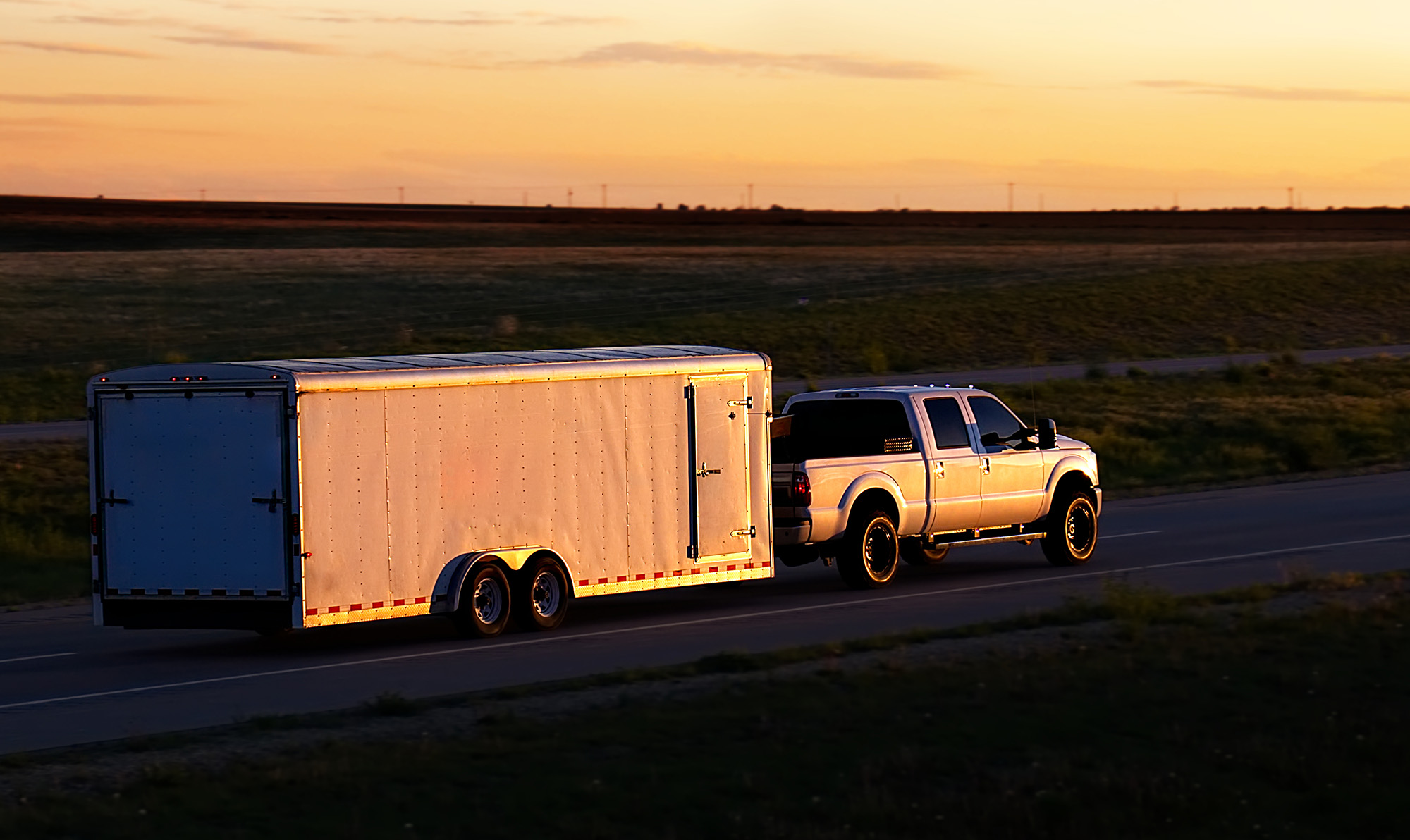 A large white pickup truck towing a covered trailer on a flat highway in the sunset.