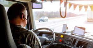 An older white male with a cellphone earpiece drives a semi-truck down a highway, with a dual-facing camera in his cab